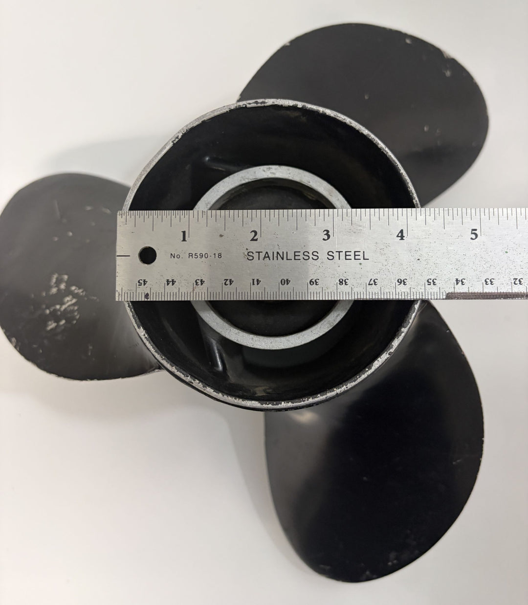 Top view of a used Volvo aluminum boat propeller with three blades and a stainless steel ruler measuring the central hub diameter against a white background.