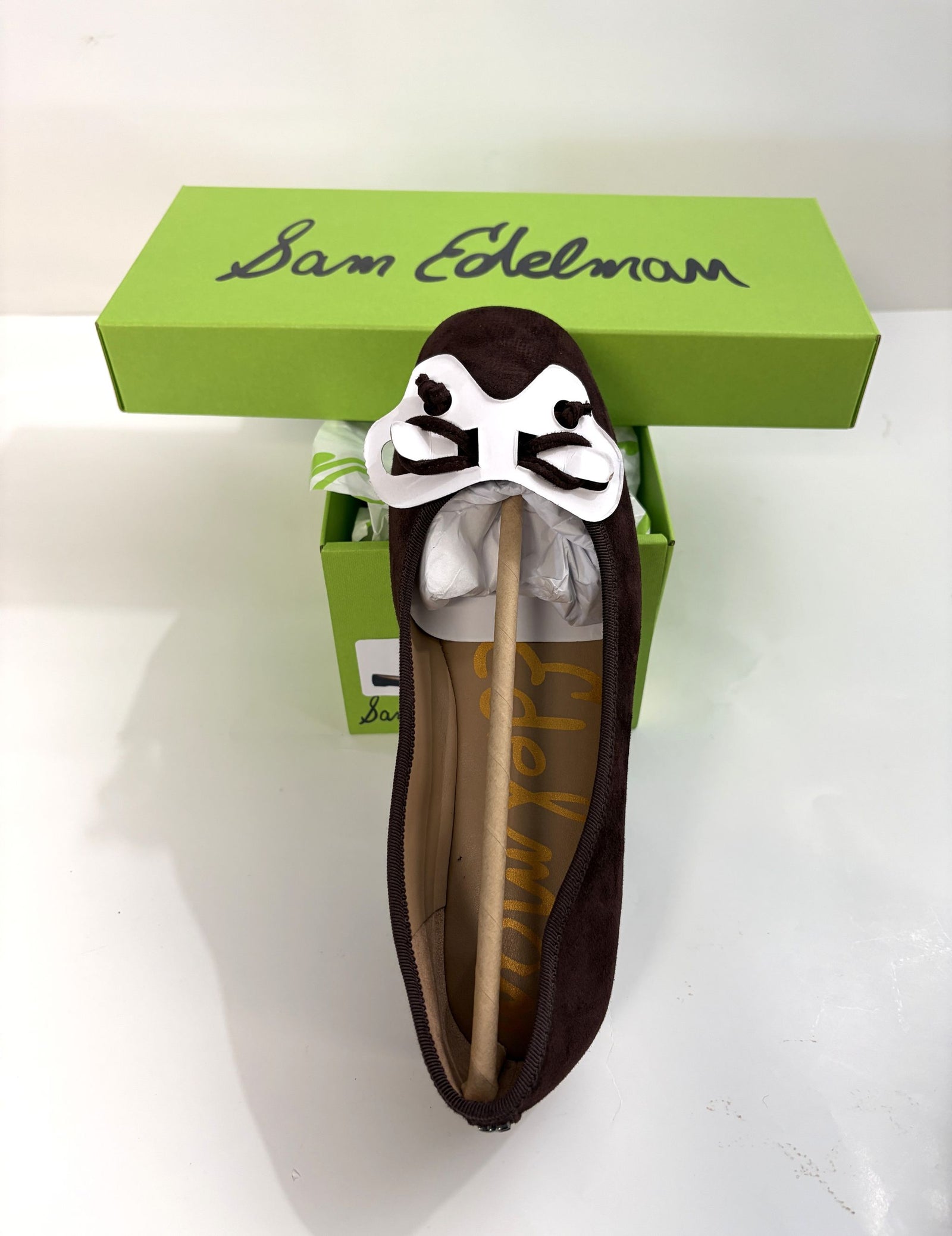 Top view of a chocolate suede ballet flat with a white decorative bow on the toe, displayed on a green Sam Edelman shoe box for casual women's footwear.