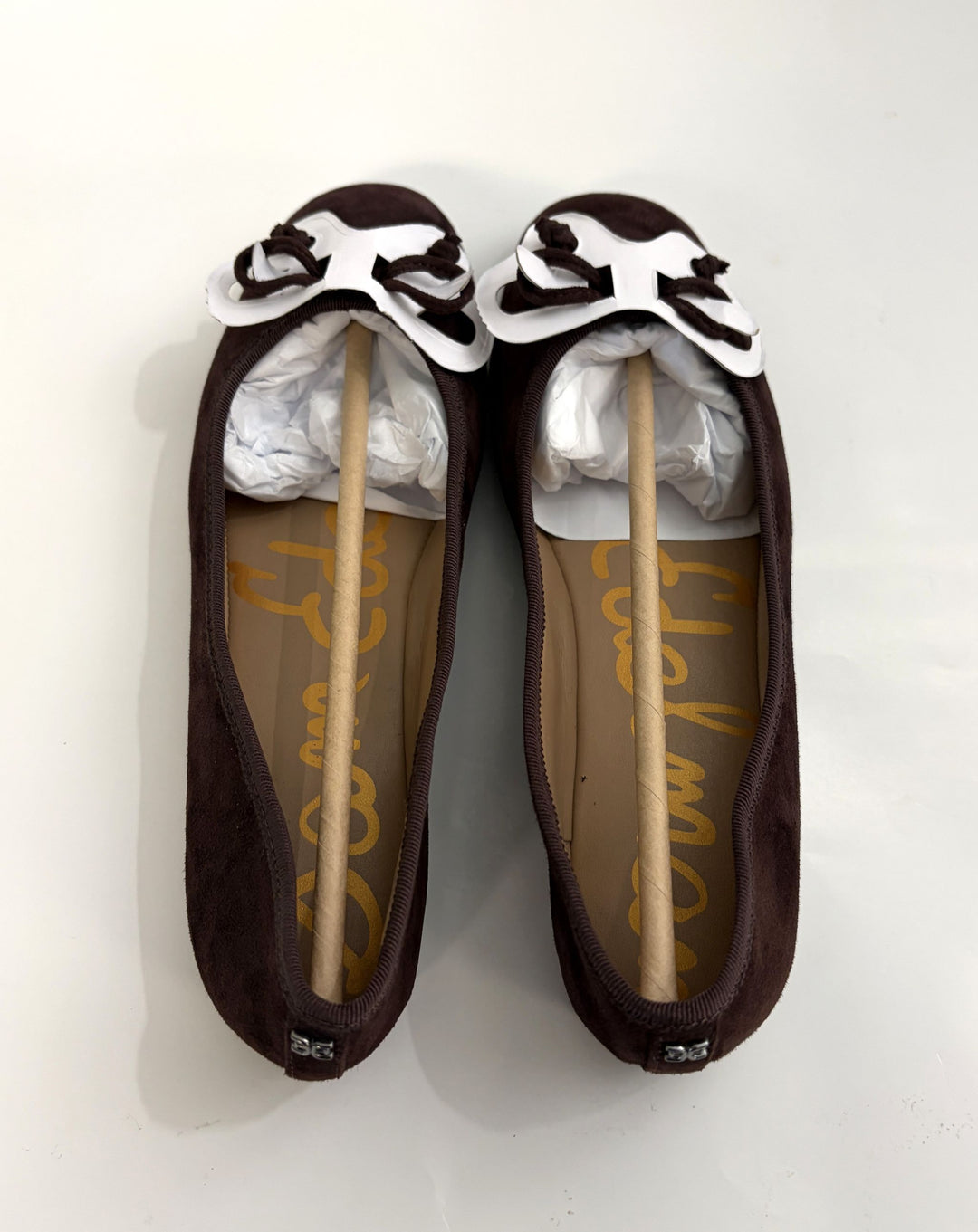 Pair of chocolate brown suede ballet flats with delicate bows on the toe, cushioned insoles, and slip-on design from Sam Edelman on a white background.