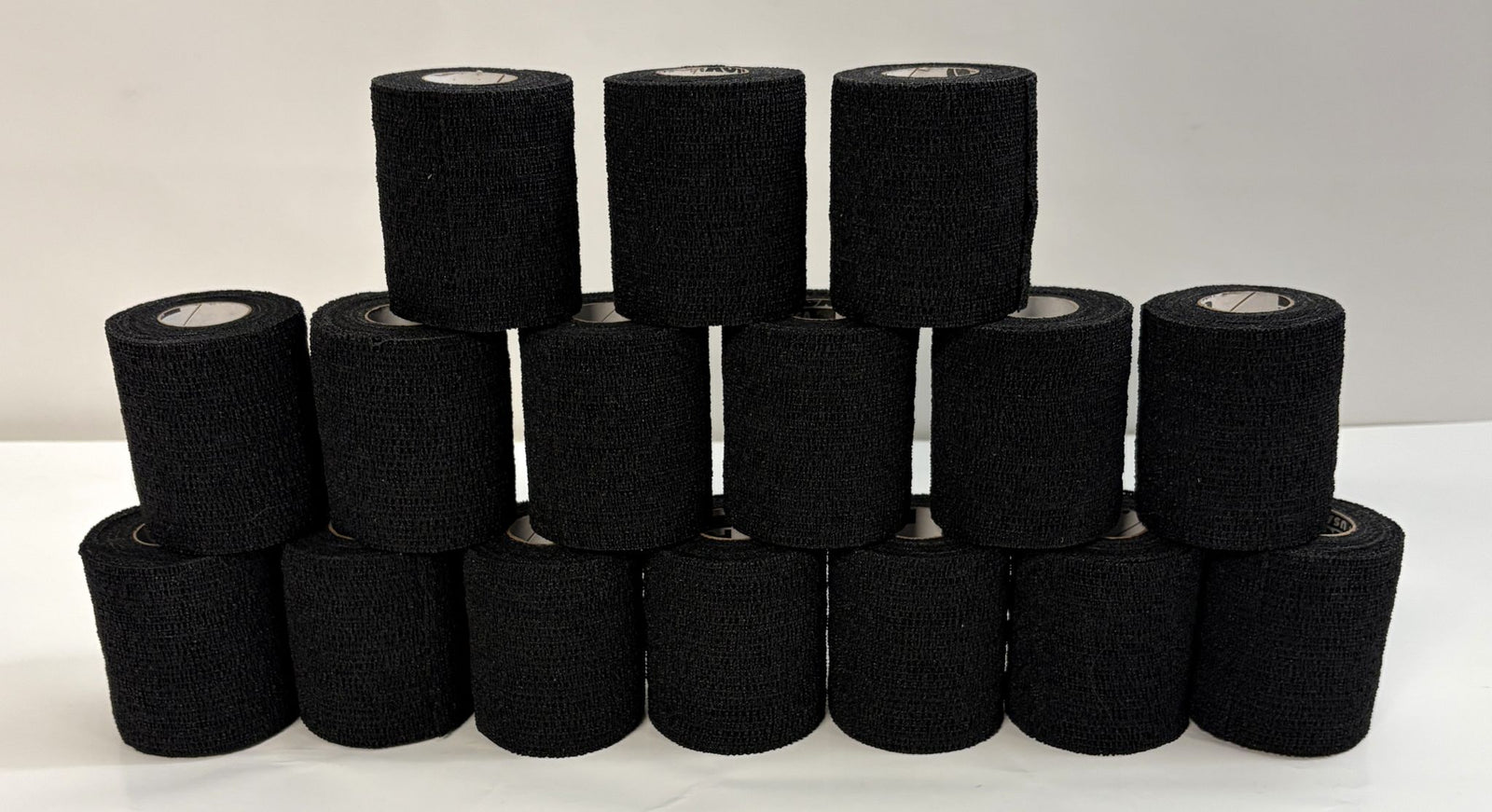 Stack of black self-adhering sports wrap rolls by Milliken, arranged in a pyramid on a white surface, suitable for athletic support and compression use.