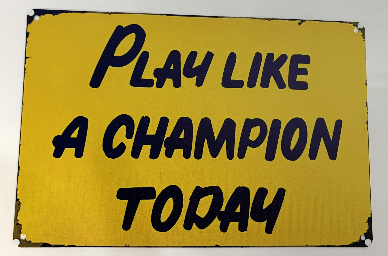 Yellow metal sign with black text that reads "Play Like A Champion Today" showing some wear and chipped paint around edges