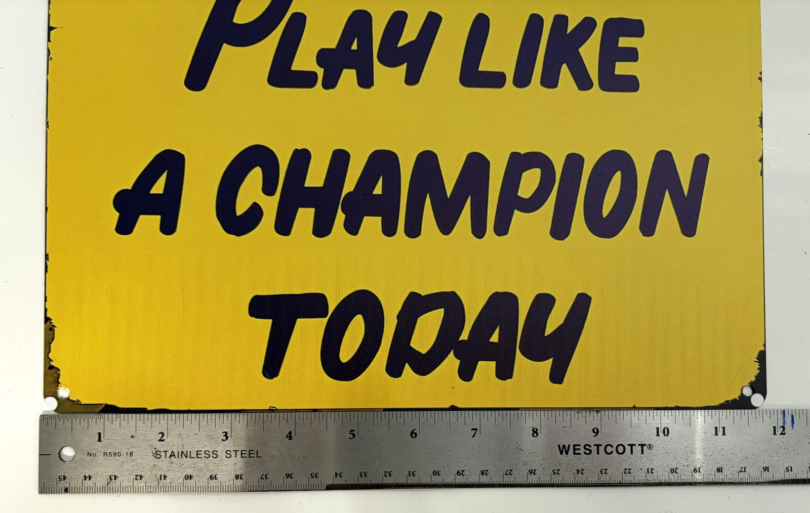 Yellow sign with black bold text reading "PLAY LIKE A CHAMPION TODAY" placed above a stainless steel ruler marked in inches.