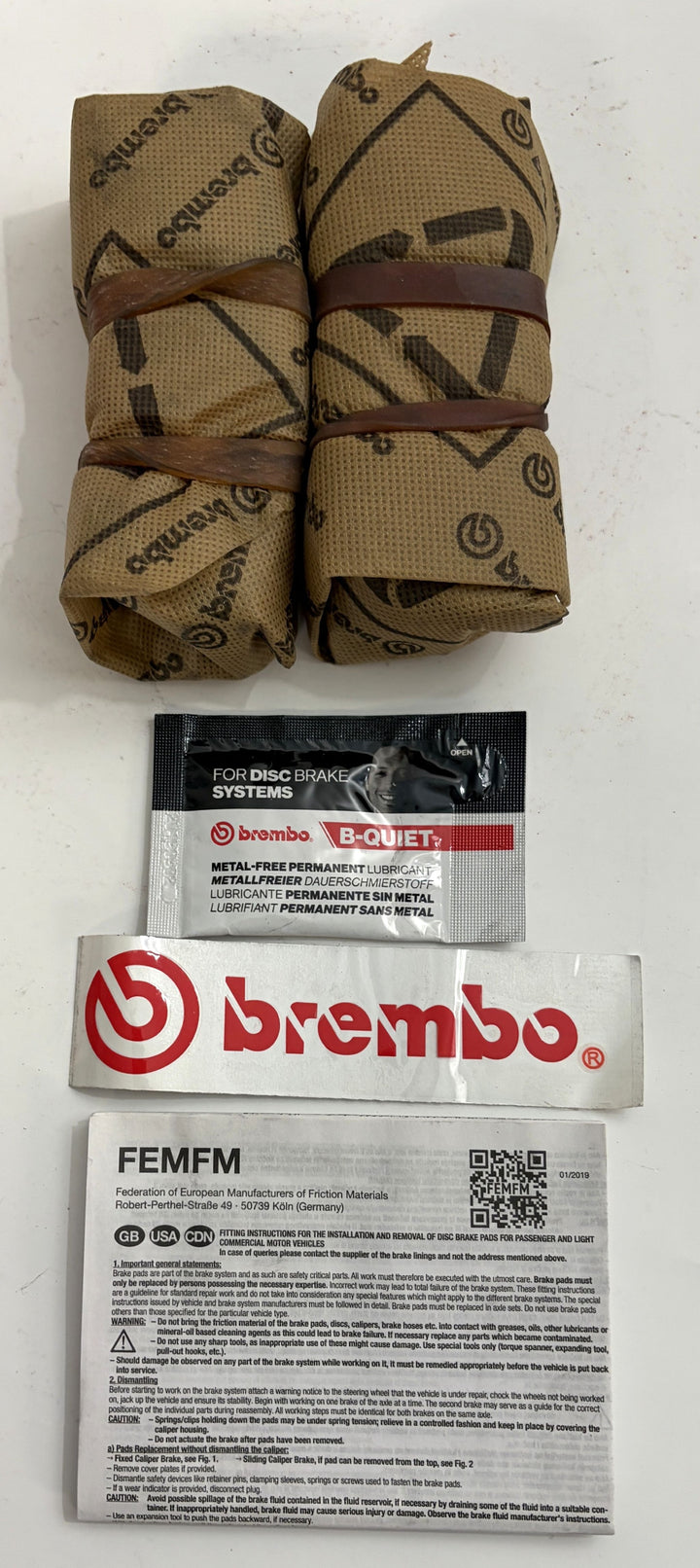 Two strapped rolls of tan Brembo B Quiet material beside Brembo labels and an instruction sheet for disc brake systems