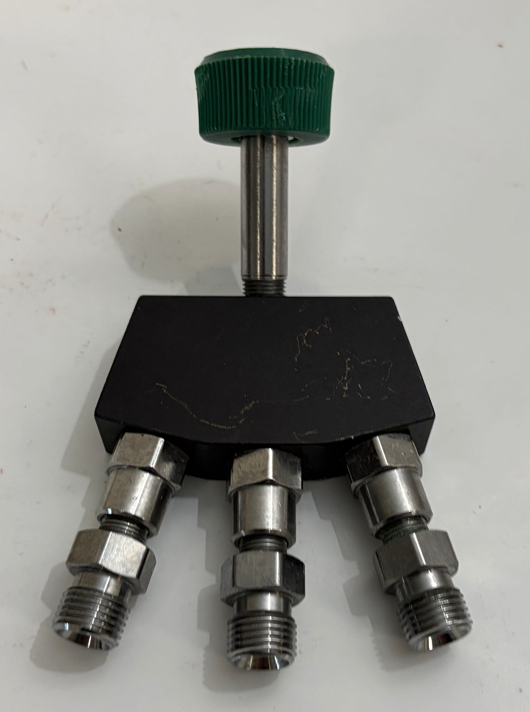 Industrial three port valve manifold with black housing, stainless steel threaded fittings, and a green adjustable hand knob on a white surface