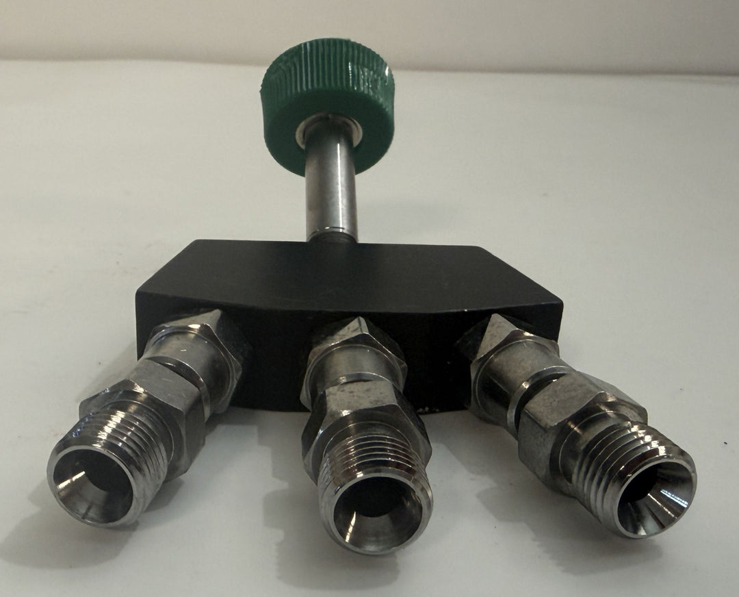 Industrial three-port metal valve manifold with threaded fittings, black rectangular body, stainless steel connections, and green handwheel.