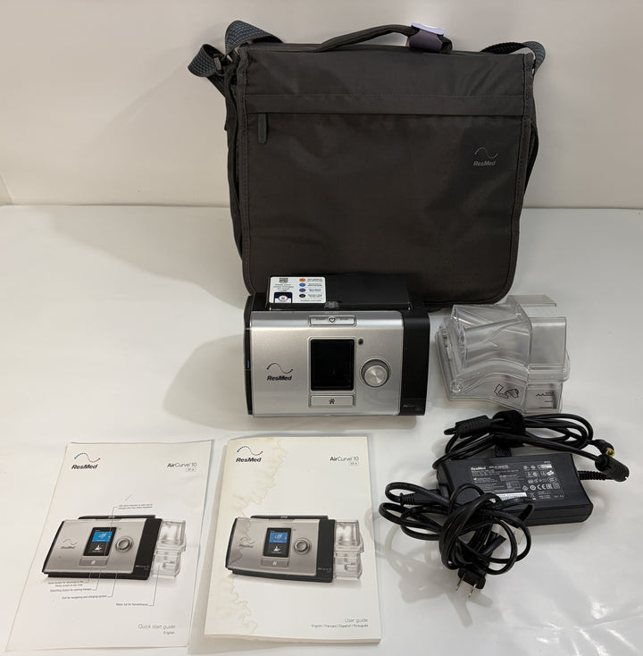 ResMed CPAP machine with black carrying case, clear humidifier chamber, power adapter, and instruction manuals arranged on a white surface