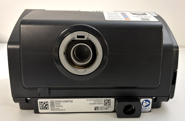 Black multimedia projector with a large central lens, control label on top, identification label below, and warning sticker on the front panel