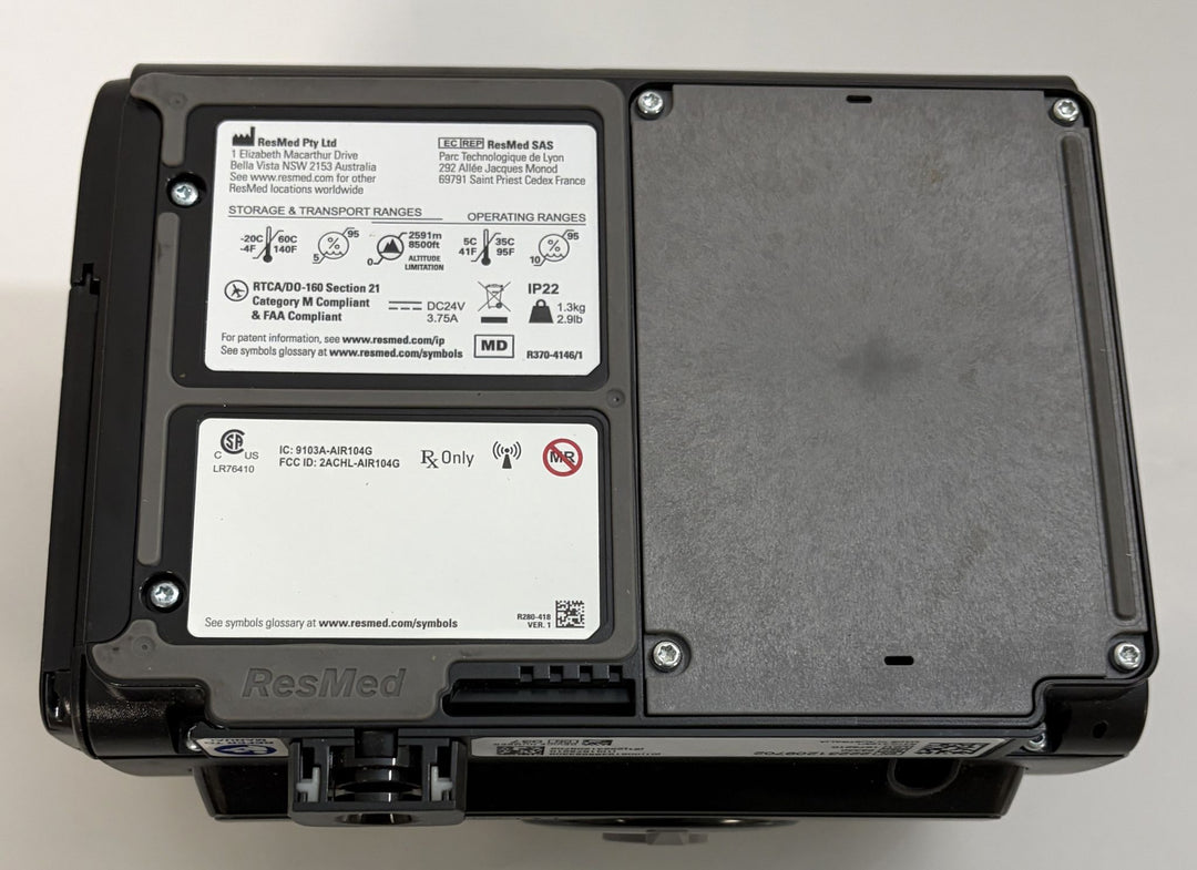 Black ResMed medical device housing with regulatory labels, a textured removable cover, mounting screws, and visible ports on the underside