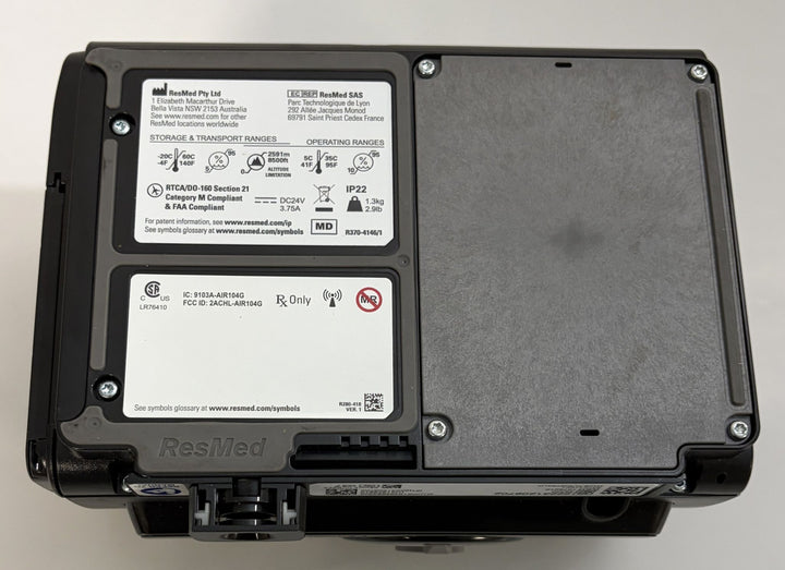 Black ResMed medical device housing with regulatory labels, a textured removable cover, mounting screws, and visible ports on the underside