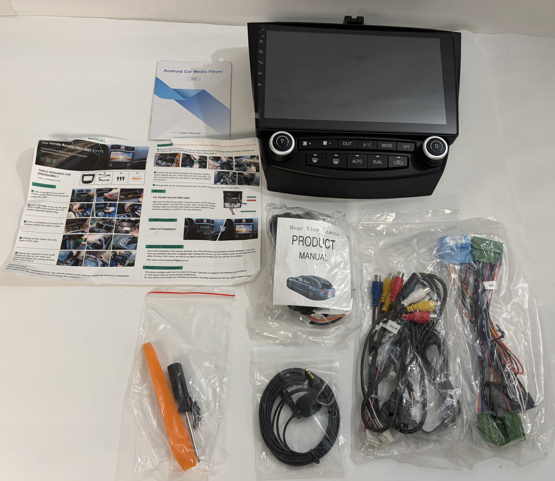 Automotive touchscreen stereo receiver with control knobs, wiring harnesses, cables, installation accessories, and manuals arranged on a white surface.