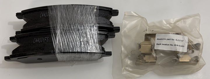 Set of Akebono ceramic brake pads stacked and wrapped in clear plastic next to sealed replacement brake pad components in branded packaging for automotive use.