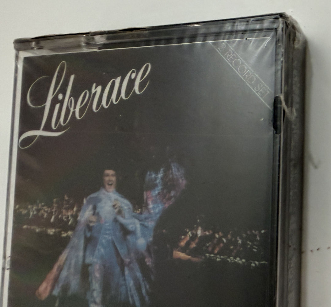 Close-up of a vintage Liberace live concert cassette in a clear plastic case, featuring bold script text and an image of the performer in an elaborate costume, from PFM/Silver Eagle Records.