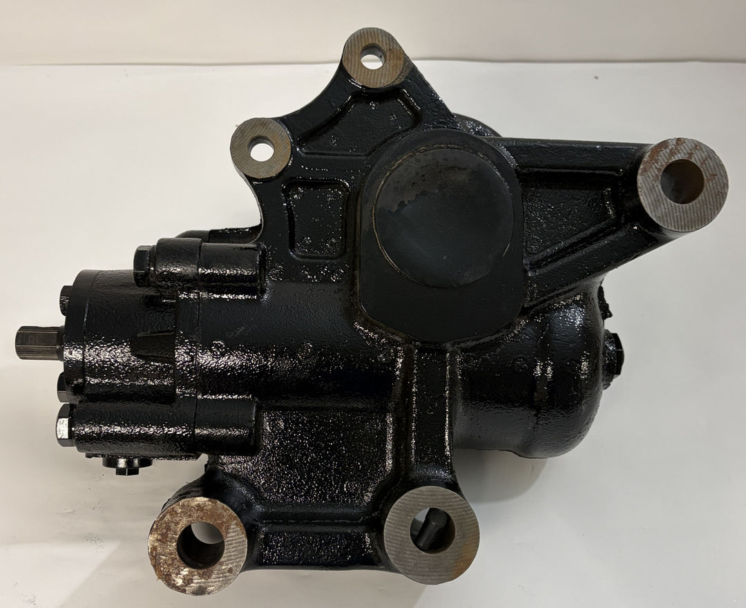 Black metal steering gear component with mounting holes and a cylindrical section, shown on a white surface, part of Sheppard’s industrial automotive parts inventory.