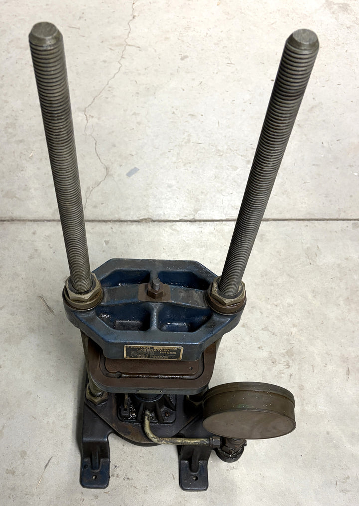 Top view of a vintage Carver Inc hydraulic press with two long threaded rods and a circular pressure gauge attached, resting on a concrete floor in an industrial setting.