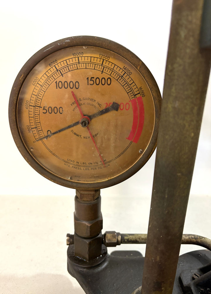 Close-up of a vintage Carver Inc pressure gauge measuring hydraulic load in pounds, attached to a metal base with signs of wear and use.