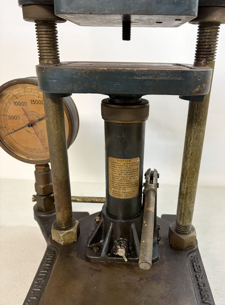 Close-up view of a vintage Carver Inc hydraulic bench press showing the pressure gauge, metal screws, and a warning label on the central cylinder in an industrial setting.