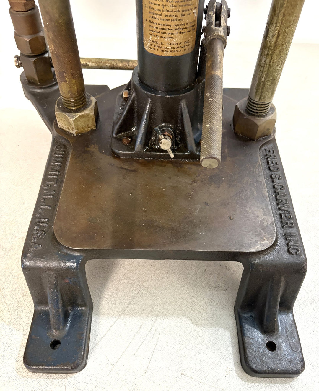 Close-up view of the base and mechanical components of a pre-owned Carver Inc hydraulic bench press showing metal construction and adjustment handle on a white surface.