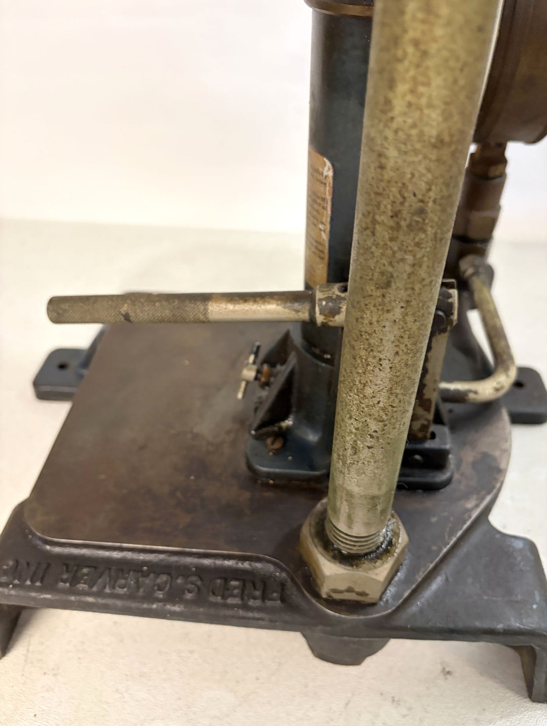 Close-up of a pre-owned Carver Inc hydraulic bench press base showing worn metal components and mounting hardware on a white surface.