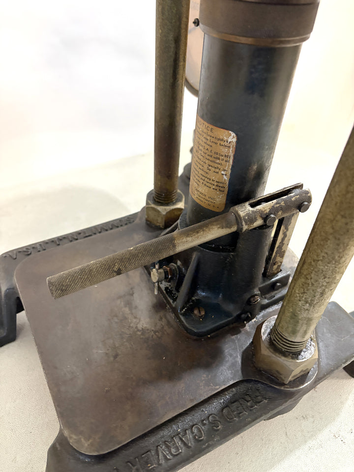 Close-up of a vintage Carver Inc hydraulic bench press showing the metal base plate, adjustment handle, and vertical support rods on a light surface.