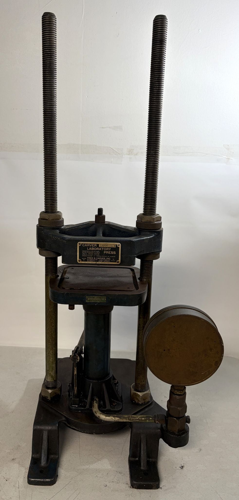 Pre-owned Carver Inc hydraulic laboratory press with vertical metal rods, pressure gauge, and sturdy black cast iron base designed for industrial and scientific use.