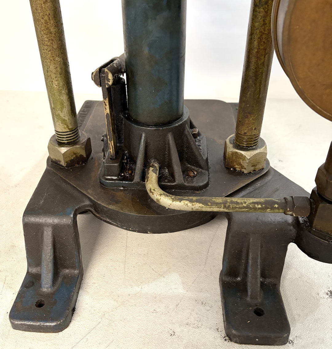 Close-up of a heavy-duty hydraulic bench press base showing metal support rods, a central blue cylinder, and connected brass fittings on a white surface, by Carver Inc.
