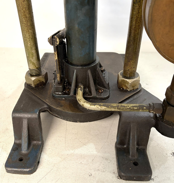 Close-up of a heavy-duty hydraulic bench press base showing metal support rods, a central blue cylinder, and connected brass fittings on a white surface, by Carver Inc.