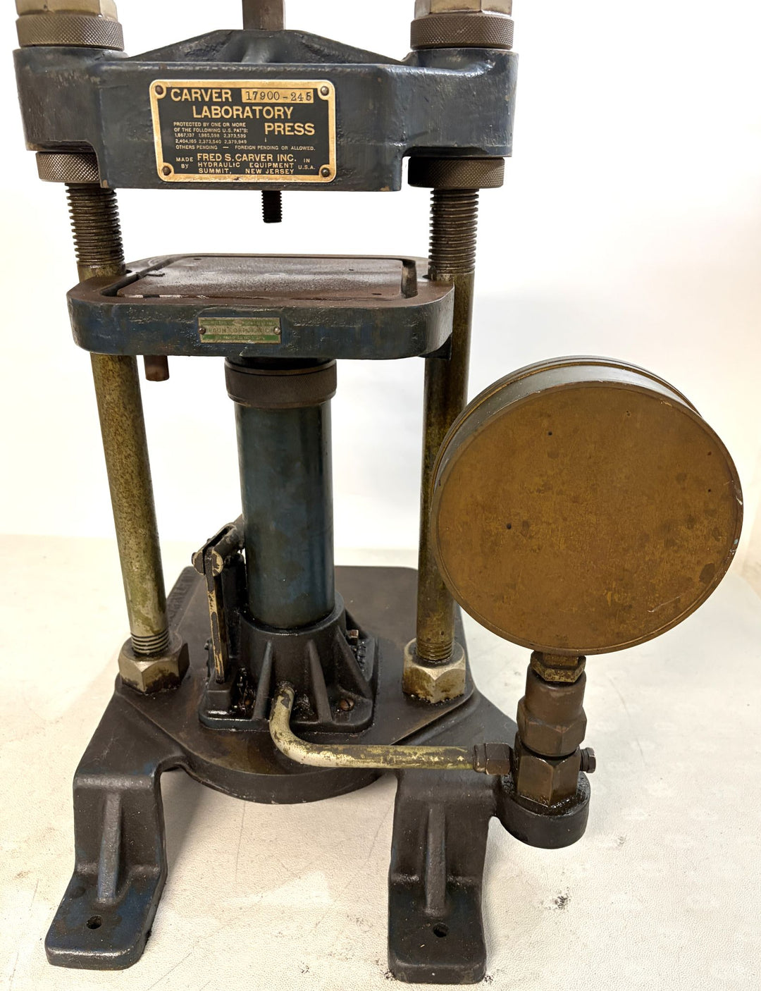 Vintage Carver Inc hydraulic laboratory press with metal base, vertical support rods, and pressure gauge, showing signs of use on a white background.