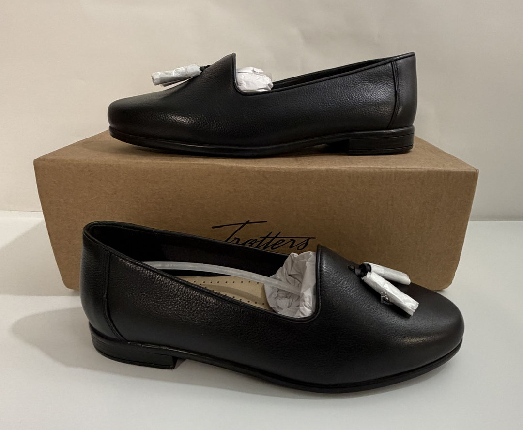 Pair of black leather tassel loafers with low heels displayed on and in front of a brown Trotters branded shoebox on a plain white surface.