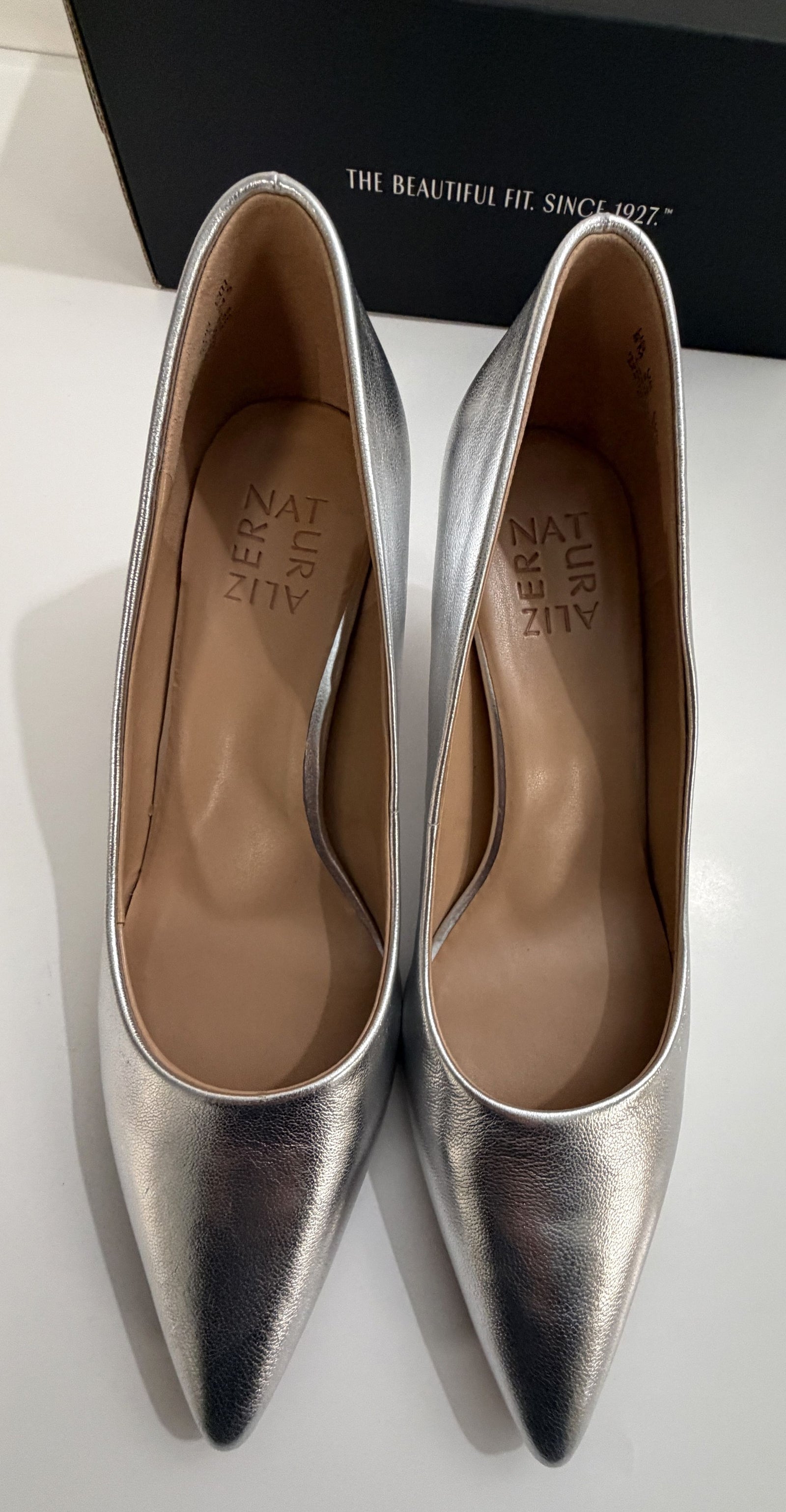 Pair of metallic silver pointed-toe women's shoes with tan insoles placed side by side on a white surface with a black box in the background.