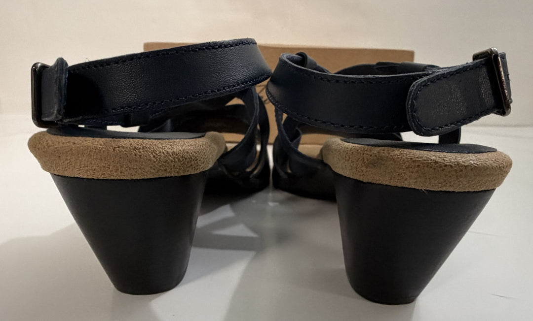 Pair of navy strappy heeled sandals with wide cushioned beige insoles and adjustable ankle straps, shown from the back on a white surface with a brown box in the background.