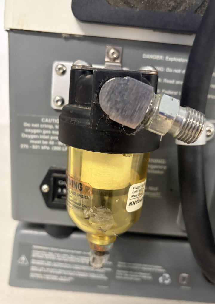 Close-up view of a transparent brass and black air or fluid filter with a metal fitting attached, mounted on industrial equipment, featuring warning labels and hoses nearby.