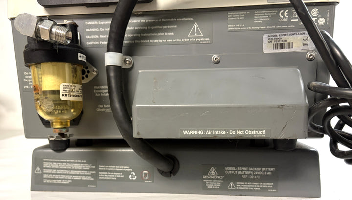 Rear view of a Philips Respironics Respironics Esprit V1000 unit showing air intake with warning label, connected tubing, and power cord on a gray metal base.