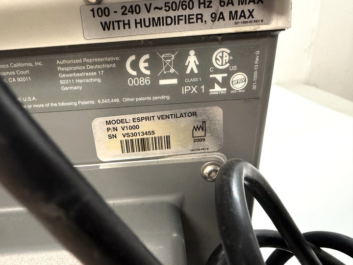 Close-up of the back panel of a Philips Respironics Esprit V1000 ventilator showing regulatory labels, model and serial number stickers, and part of a black power cable.