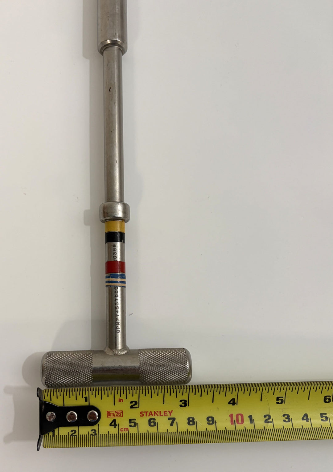 Close-up of a metal T-handle hex driver with colored bands on the shaft, placed beside a yellow Stanley measuring tape showing size in inches, from PHENTERSALES.