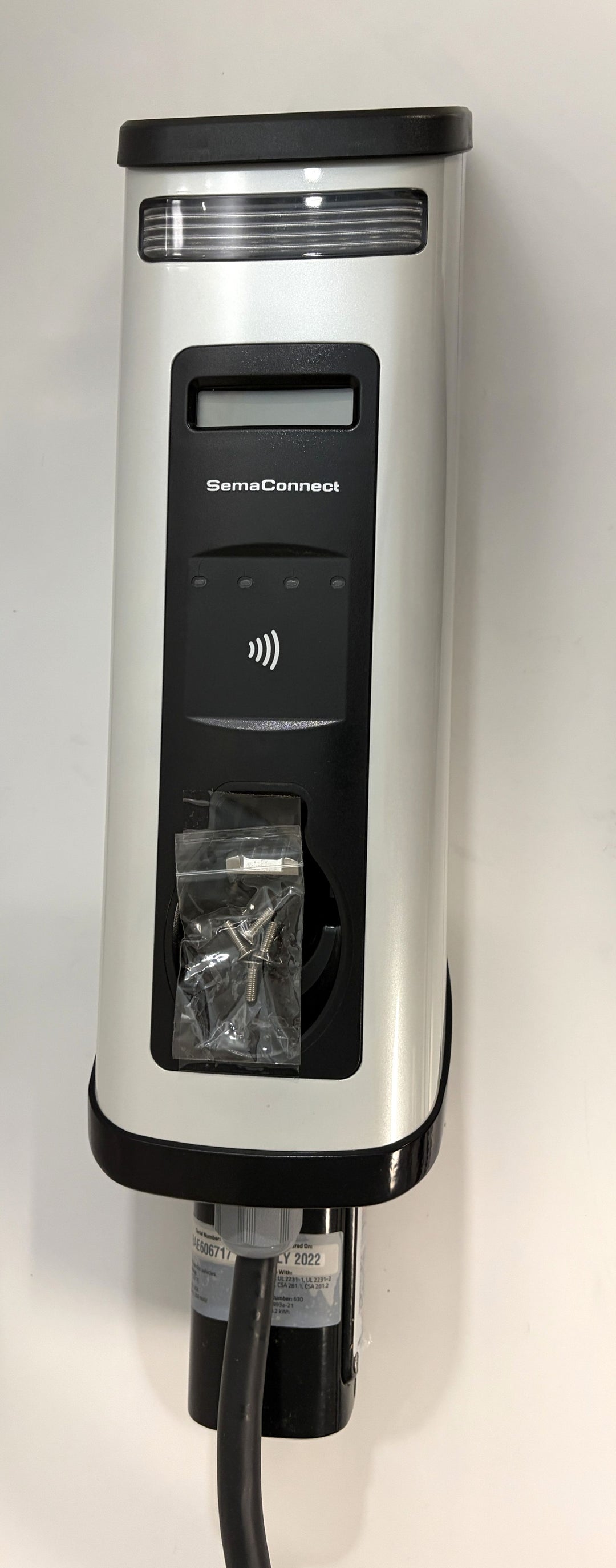 Vertical view of a SemaConnect commercial-grade EV charging station in silver and black with attached power cable and small hardware packet on a white background.