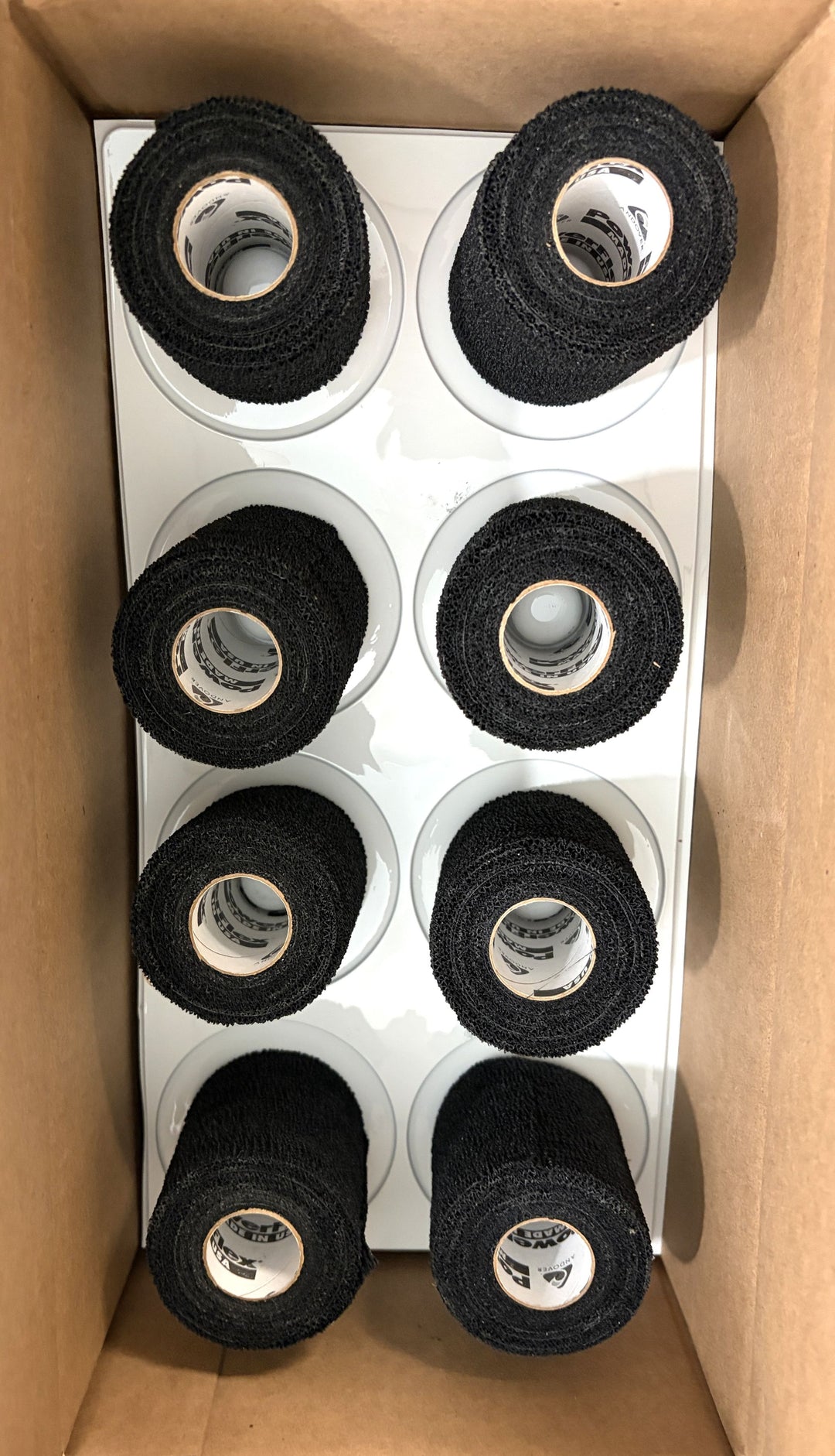 Eight black sports wrap rolls neatly arranged inside a cardboard box, featuring textured fabric and white inner cores, suitable for athletic or medical use by Milliken.