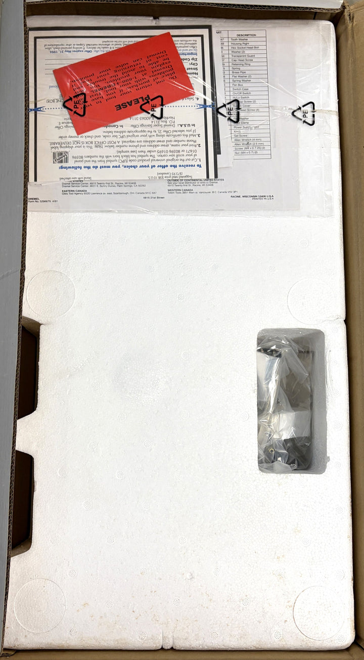 Top view of a Dremel scroll saw partially covered by molded protective foam inside an open shipping box, with accompanying paperwork and a red plastic warning label included.