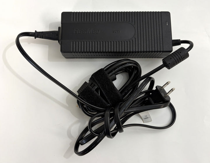 Black Resmed 90W power adapter with attached cable and two-prong plug, shown coiled and secured with a Velcro strap on a white surface.