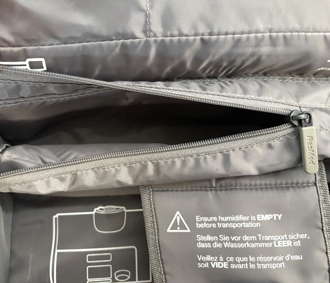 Interior view of a charcoal gray Resmed travel bag showing a partially opened zippered compartment and printed multi-language instructions to ensure the humidifier is empty before transport.