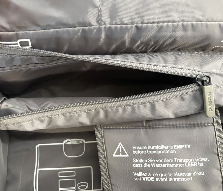 Interior view of a charcoal gray Resmed travel bag showing a partially opened zippered compartment and printed multi-language instructions to ensure the humidifier is empty before transport.