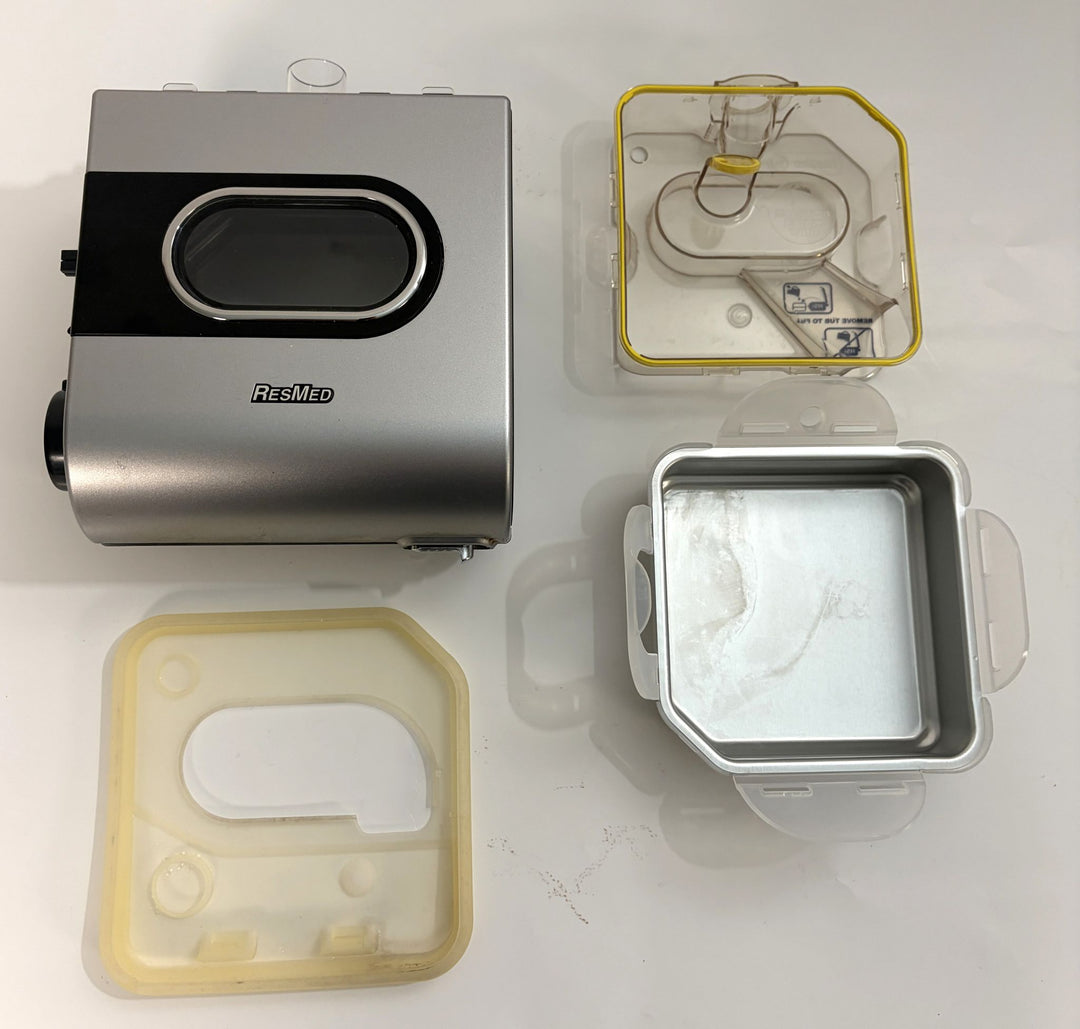 Silver and black ResMed humidifier unit alongside three clear and frosted plastic humidifier components with yellow sealing edges, shown on a white surface.
