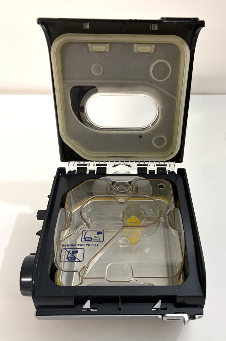 Open pre-owned Resmed humidifier reservoir component showing clear water chamber and hinged black plastic lid with filling instructions and yellow valve detail inside.