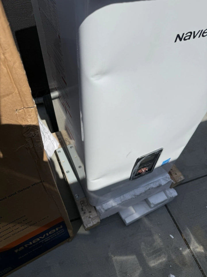 White Navien appliance with a gray base and front control panel, positioned beside a cardboard shipping box on a concrete surface