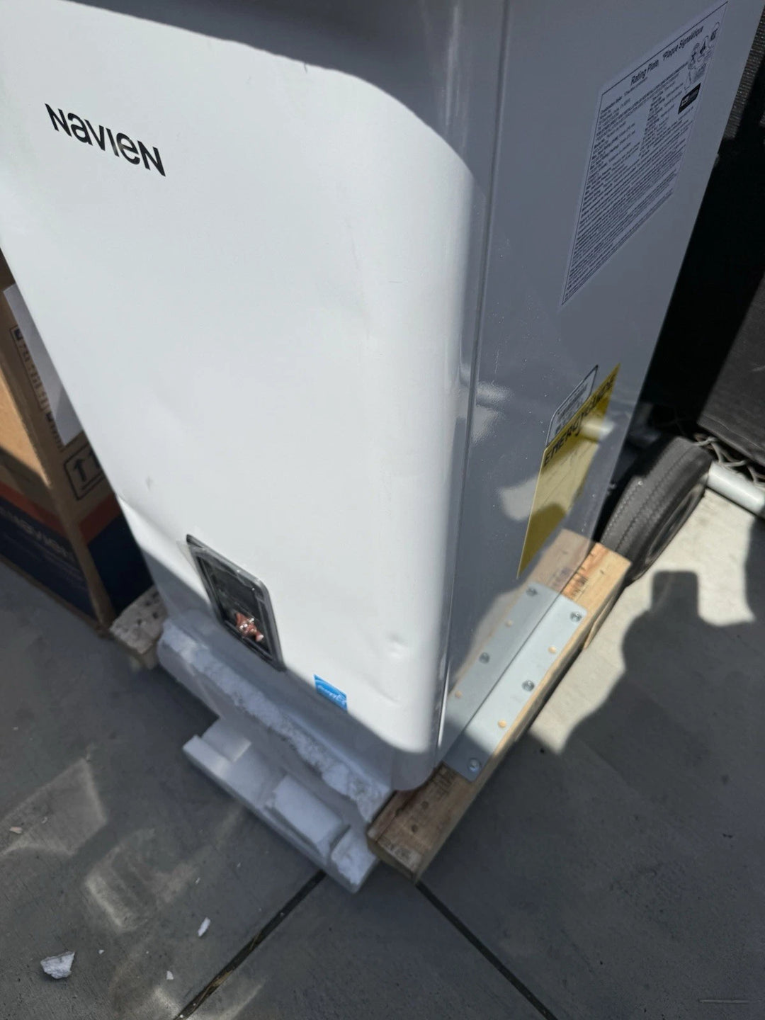 White Navien heating appliance with identification label, mounted on a wooden pallet outdoors.