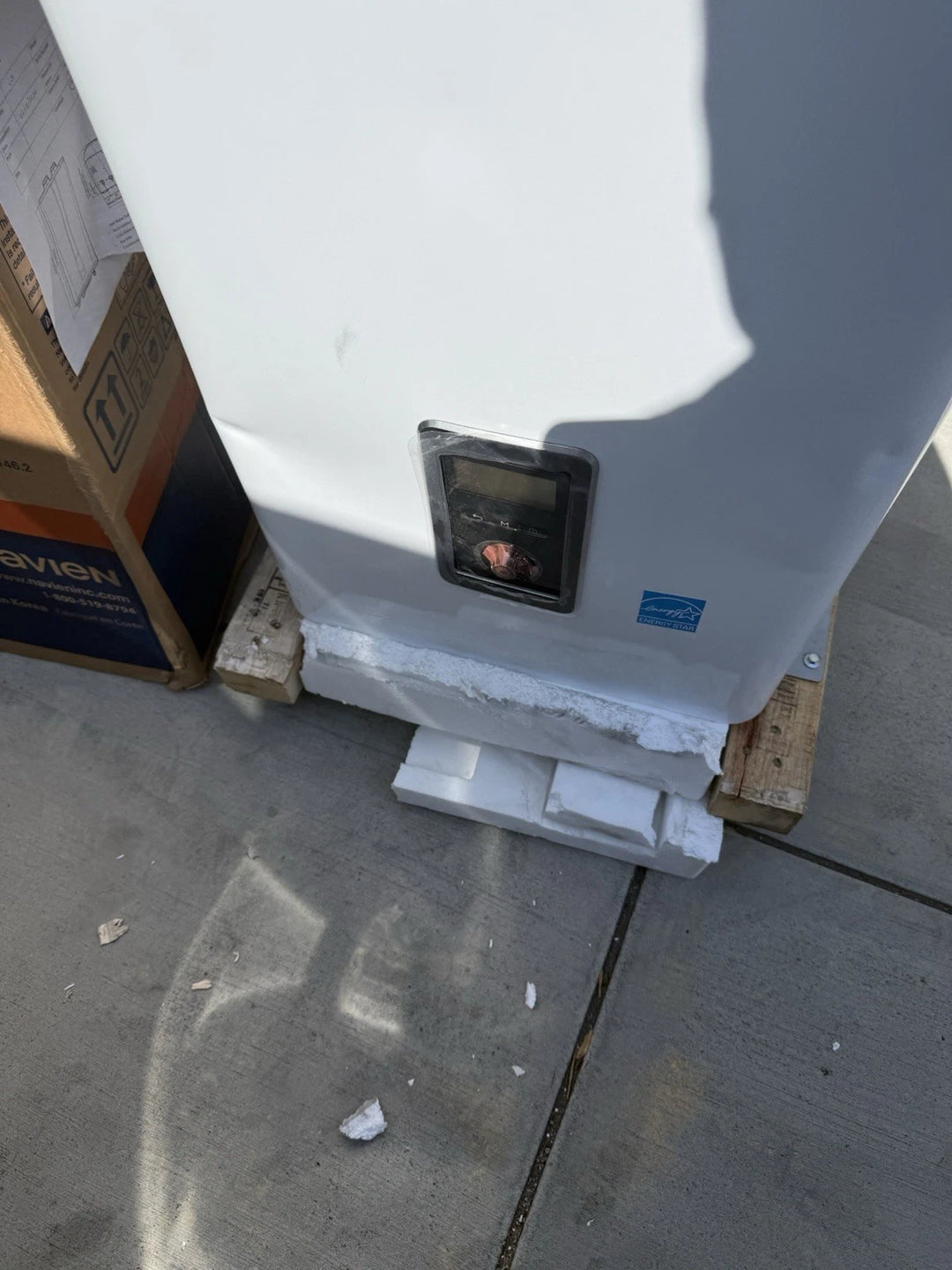 White industrial equipment with a black rectangular inset panel and blue label, secured with protective packaging on a wooden pallet outdoors